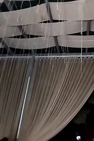 A close-up shot of a decorative canopy made of white fabric strips and strung with lights. A large beige curtain hangs from the bottom of the canopy, which appears to be outdoors at night.