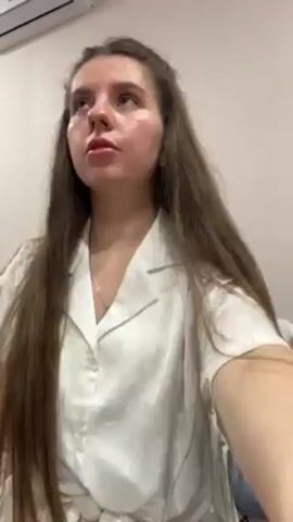 A young woman with long brown hair, wearing a white button-down shirt, stands in front of a white wall with a white air conditioner mounted near the ceiling. She holds a clear glass filled with a red liquid, possibly juice or a cocktail. She takes a sip from the glass and then lowers it. Music plays audibly in the background.