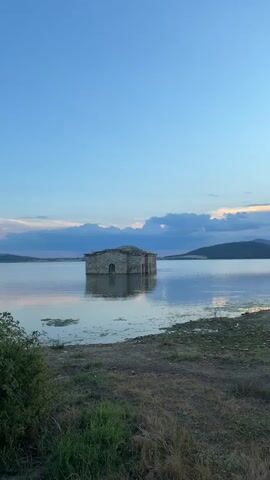 The image shows a submerged stone church in the middle of a lake under a twilight sky. The church is made of stone and has a traditional architectural style with a rounded roof and arched windows. It is partially submerged in the water, with only the upper portion of the structure visible above the surface. The lake is calm and reflects the sky and the church. The sky is a gradient of blue and purple, with scattered clouds tinged with pink and orange from the setting sun. In the foreground, there is dry grass and some sparse vegetation along the shoreline. The overall scene is serene and somewhat melancholic, evoking a sense of history and abandonment. The time of day is dusk, and the weather appears to be clear with some cloud cover. The location is a lake in Panicherevo, Bulgaria, where the church is a notable landmark, often referred to as the "Sunken Church of Zhrebchevo." There is no visible text in the image.