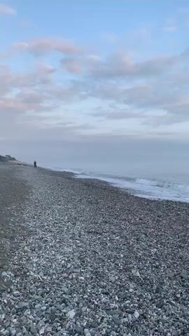 The video shows a static shot of a pebble beach meeting the ocean under a partly cloudy sky. The beach is covered with gray pebbles. The water is calm with small waves washing onto the shore. The sky is light blue with scattered white and gray clouds, some tinged with pink. The horizon line is visible, though somewhat obscured by a slight mist. The overall impression is of a quiet, serene coastal scene.
