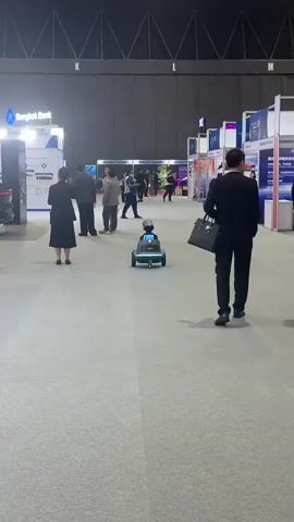 A mobile robot moves across a convention center floor. The robot is black and gray with a blue screen displaying a red symbol. It has a light strip along the bottom that is illuminated. Several people are visible in the background, some walking and some standing and talking. Booths with displays are set up on either side of the aisle. The floor is carpeted in a light gray. The lighting is artificial.