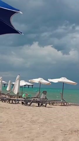 The video shows an empty beach with lounge chairs and umbrellas set up facing the ocean. The sky is overcast with dark clouds, suggesting potential rain or a storm. The water is a clear turquoise color, with small waves breaking on the shore. A pier or structure extends into the water in the distance. The overall impression is of a beach on a cloudy day, possibly before or after a storm.