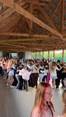 The video shows a group of young people dancing in a circle inside a wooden pavilion. The ceiling is made of exposed wooden beams and the walls appear to be open to the outdoors. The subjects are a mix of boys and girls, all appearing to be teenagers, who are holding onto each other's shoulders as they move in a circular dance. They are dressed in casual, modern clothing.

In the background, there are more people gathered, some of whom appear to be standing and watching, while others might be part of the dance as well, further down the line. There is a stage or platform in the distance with a screen and possibly a DJ or speaker. The lighting suggests it is daytime, possibly late afternoon or early evening, with sunlight filtering in from the sides. The overall atmosphere seems to be energetic and celebratory, with a sense of community and fun.

One notable detail is the young girl in the foreground, closer to the camera, who is looking to the side and has dyed red streaks in her hair. She is wearing glasses and a light pink shirt, and her expression is neutral.

The text visible on the screen in the background appears to be "YOUTH." The setting, with its open-air pavilion and natural surroundings visible through the openings, suggests an outdoor event or gathering, possibly at a camp or a recreational area. The people's clothing and hairstyles are consistent with contemporary fashion. The activity is clearly a group dance, likely to music being played.