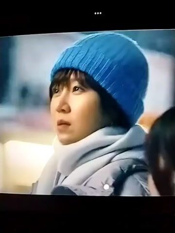 The video shows a close-up of a young woman wearing a blue beanie and a light grey scarf, looking off-camera to her left. She has dark hair and bangs. Subtitles are visible at the bottom of the screen. The background is blurred, suggesting an outdoor or dimly lit setting with some lights visible. The camera then slightly pans to the right, showing another person's profile in the foreground, also looking to their right. The subtitle text remains visible, and the woman in the foreground looks directly at the viewer with a questioning expression. The text then changes. The scene appears to be from a drama or movie, with a focus on dialogue.