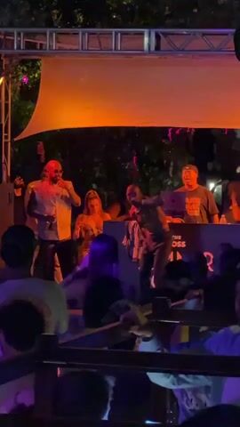 The video shows a live outdoor music performance at night. A stage is set up with a simple frame and a white canopy, illuminated by orange and purple lights. Four men are visible on the stage: two stand at the front, one is at a DJ setup, and another stands beside it. The men at the front appear to be singing or rapping into microphones. The man on the right is shirtless. In front of the stage, a crowd of people is gathered, watching and filming the performance with their phones. A wooden railing separates the crowd from the stage. A sign with the words "SUMMER" and "THE BOSS" is visible in the foreground, behind the railing. Trees are visible in the background.