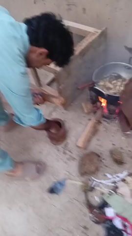 A person is shown bending over a mortar and pestle, preparing to grind something. In the background, a cooking pot is simmering over an open fire. The ground is dusty and uneven. The person is wearing a light blue shirt and light-colored pants, and appears to be wearing slippers. There is debris on the ground, including what looks like scraps of fabric and some sort of dried plant material. The overall scene suggests an outdoor cooking environment.