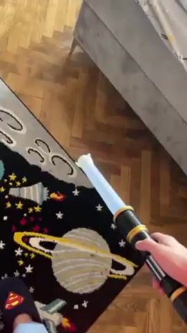 From a high-angle perspective, a hand holds a toy lightsaber with a black and gold hilt. The lightsaber is activated, emitting a bright blue light. The hand is wearing a Superman-themed slipper. The lightsaber is held above a rug featuring a space theme with planets, stars, and rockets.  A portion of a gray couch is visible in the background, and the floor is parquet wood.