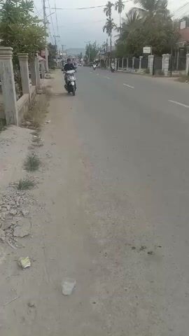 A person is riding a white scooter on a dirt road next to a paved road. The person is wearing a dark jacket and jeans. There are trees and buildings on the side of the paved road. The sky is overcast. A second scooter with two people on it passes by in the background.
