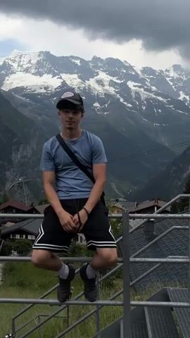 This video features a young person posing for a photo with a majestic mountain landscape in the background. The subject, a male with dark hair, is wearing a blue t-shirt, black shorts with white stripes, a baseball cap, and a black crossbody bag. He is sitting on a metal railing, looking directly at the camera with a neutral expression.

The setting is a mountainous region, with snow-capped peaks dominating the horizon under a cloudy sky. Below the mountains, a village with traditional Swiss chalets is nestled in the valley, with trees and greenery adding to the picturesque scene. A ski lift cable is visible in the distance, suggesting this location is popular for outdoor activities. The overall atmosphere is serene and awe-inspiring, conveying a sense of adventure and natural beauty.

The weather appears overcast, with dramatic clouds adding to the intensity of the mountain scenery. The time of day is likely daytime, given the ambient light. The location cues strongly suggest Mürren, Switzerland, known for its stunning alpine views. There is no visible text in the media file.