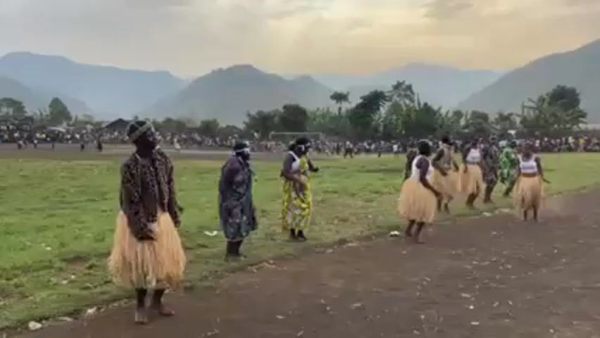 A group of seven people are dancing in a field in what appears to be a public performance. The group consists of women wearing colorful tops with white tank tops underneath and light brown, grass-like skirts, and one man wearing a leopard print jacket over a white tank top and a similar skirt. The dancers are in a line near a dirt track or path on the edge of a large grassy field. A crowd of people is standing in the background, suggesting an audience. In the distance, there are lush green mountains under a cloudy sky. A soccer goal can also be seen on the field. The overall setting suggests a rural or semi-rural area.
