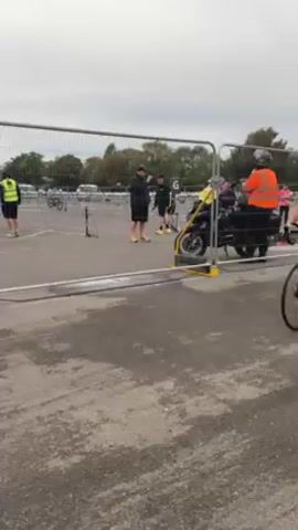 The video shows a cyclist riding a black bicycle on a wet, gray asphalt surface. The cyclist is wearing a blue and white athletic outfit with a helmet.  He is passing by a metal barrier. In the background, there are parked vehicles, including a white van. Several people are also visible, including a person in a yellow safety vest and another in an orange vest. A person stands near the camera, holding up a mobile phone to capture a photo or video of the cyclist. The phone screen displays the time, 8:57. The overall environment appears to be an outdoor event or race.