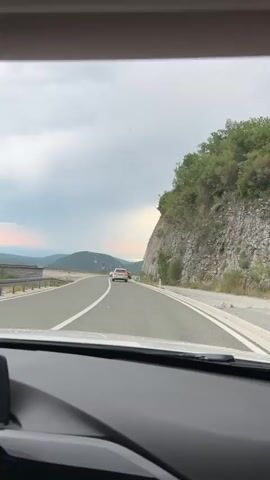 The video captures a first-person view from inside a vehicle traveling on a paved road. The road curves to the right, bordered by a steep, rocky cliff face on the right and a guardrail on the left, beyond which lies a valley with mountains in the distance. The sky is overcast with patches of sunlight breaking through, casting rays onto the landscape. Several cars are visible on the road ahead, including two white cars in front of the viewpoint, and further ahead, a white SUV. The environment suggests a scenic route, possibly coastal or mountainous, with vegetation clinging to the cliff and hillsides. The footage is shot in a vertical orientation.