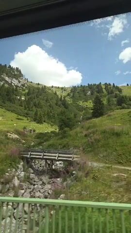 The video captures a view from a moving vehicle, likely a bus or car, looking out a window. The scene is a mountainous landscape with lush green slopes covered in trees and grass. A rocky, rugged mountain peak is visible in the background under a bright blue sky with scattered white clouds. A winding path or road is visible in the lower left of the frame. There are some scattered rocks and vegetation along the roadside. A small wooden structure, possibly a mailbox or information sign, stands on a post in a grassy area. In the distance, on the right side of the frame, there appears to be a small bridge or crossing over a stream. The foreground features a green railing, suggesting the viewer is inside a vehicle with a window and railing. The lighting indicates it is daytime, likely late morning or afternoon. The overall impression is one of a scenic drive through a natural, mountainous environment.
