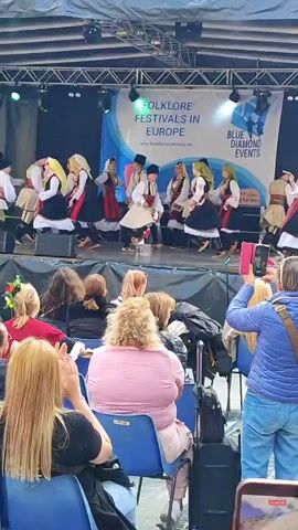 The video shows a group of people performing a folk dance on a stage labeled "Folklore Festivals in Europe" in front of an audience. The dancers are dressed in traditional costumes with white tops and black bottoms, accented by red and white waist sashes and decorative headpieces. The men are wearing black hats, and the women have their hair arranged with various decorations.

The dancers are moving in synchronized steps, lifting their arms and swirling as they perform. Some members of the audience are watching attentively, while others are recording the performance with their phones. The audience members are seated in blue plastic chairs. Luggage and personal items are scattered around the seats. The lighting on the stage is bright, with spotlights shining on the dancers. The stage is covered with a green carpet, and the backdrop includes a logo for "Blue Diamond Events".