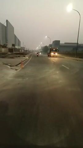 The video shows a road with several motorcycles. In the foreground, the road is visible with lane markings. A truck is approaching from the opposite direction. Further down the road, multiple motorcycles are traveling in the same direction as the camera. The environment appears to be an industrial area with large buildings on either side of the road. The sky is hazy or overcast, and streetlights are illuminating the road. The overall impression is of a commute or travel on a highway in an urban or industrial setting, possibly during dawn or dusk.