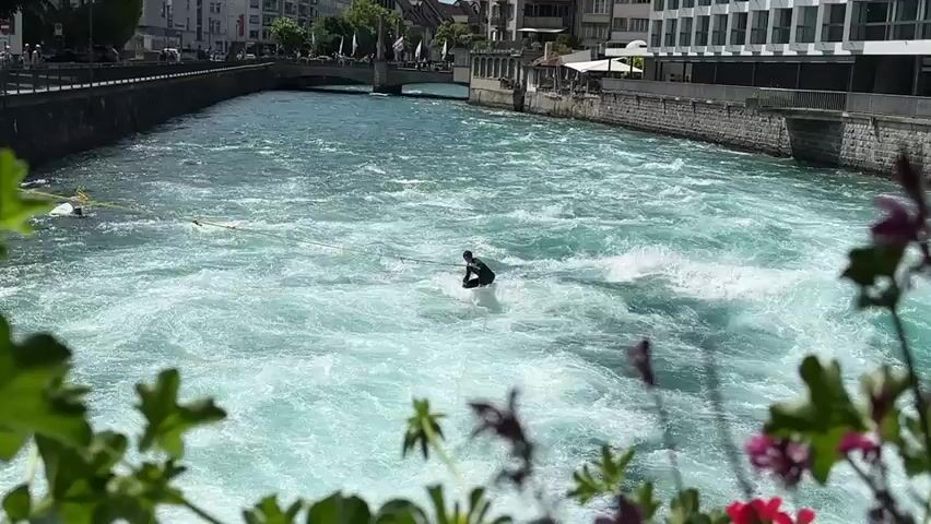 A person is wakeboarding in a fast-moving river with white water rapids. The person is wearing a dark wetsuit and riding a white wakeboard. They are attached to a cable that extends to the left of the frame, suggesting they are being towed by a cable system. The river is surrounded by a city with buildings and a bridge visible in the background. In the foreground, there are out-of-focus pink and red flowers and green leaves. The water is a bright turquoise color and appears turbulent. The sky is clear and the scene is brightly lit, indicating it is daytime.