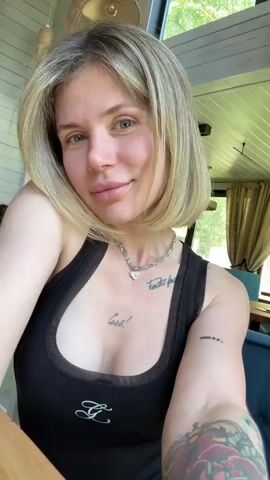 A young woman with shoulder-length blonde hair, styled in a bob with bangs, is featured in a close-up selfie video. She has light-colored eyes, fair skin, and appears to be wearing minimal or natural makeup. She is dressed in a black tank top with a sheer mesh detail at the neckline, featuring a small white stylized letter 'G' or similar logo on the front.

A silver chain necklace with a small heart-shaped pendant hangs around her neck. She has several visible tattoos: a script tattoo on her upper chest that partially reads "Cara", another script tattoo on her right upper chest/shoulder area, and a colorful tattoo partially visible on her left forearm.

The woman looks directly at the camera, occasionally shifting her gaze and offering a subtle, calm smile. The setting is indoors, appearing to be a bright room with a light-colored wooden paneled ceiling and walls. A large window in the background shows vibrant green foliage outside, suggesting a natural, possibly treed, environment. A light fixture made of natural material hangs from the ceiling. Dark curtains are visible further back in the room. The scene is well-lit by natural light, indicating it is daytime.