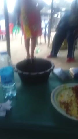 The video shows a view from under a tin roofed structure, likely a restaurant or bar. In the foreground, a table holds a black bucket, a phone, and a plate of food. Beyond the table, several people are visible. One person in a colorful dress is standing. Another person, wearing dark pants and red shoes, is leaning against a blue plastic chair. In the background, there is a table with seated people, all of whom appear to be male and dressed in white. The outdoor setting consists of a sandy ground with trees visible through the openings in the structure. The video is somewhat shaky and blurry.
