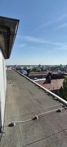 The video shows a view from a rooftop looking out over a city. In the foreground, there is a gray, gravelly rooftop with white wires and supports running along it. A white gutter runs along the edge of the building. In the background, there is a cityscape with numerous buildings, some with red tile roofs. Trees are visible throughout the city. Several construction cranes are present in the distance. The sky is blue with some thin white clouds, and the sun is shining brightly. The video appears to be a panning shot, moving from left to right.