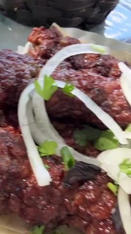 The video shows a close-up view of a plate of grilled meat, likely kebabs, garnished with sliced onions and fresh herbs, possibly cilantro and dill. The meat is dark brown and appears to be seasoned and well-cooked, served on a white platter with a rippled edge. In the background, a small glass bowl contains a red sauce, possibly tomato-based. The setting appears to be outdoors, with a marbled surface visible beneath the platter and a blurred background suggesting natural elements like stone and possibly greenery. The lighting suggests it is daytime, with sunlight casting shadows.