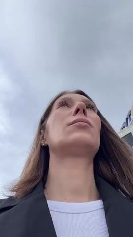 A low-angle shot of a woman looking upwards at a cloudy sky. She has long brown hair and is wearing a black jacket over a white shirt. Part of a building with purple and yellow accents is visible on the right side of the frame. The camera is angled upwards, capturing her face and the sky.