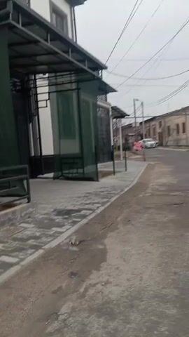 The video shows a view from a sidewalk or the edge of a street, moving slowly forward. The street is paved and has some cracks and patches.  To the left is a sidewalk with gray paving stones and a covered structure that appears to be a row of small shops or stalls, possibly closed. Across the street are older, single-story buildings with light-colored facades. A white car is parked further down the street. A person walks across the street in the distance. The sky is overcast.