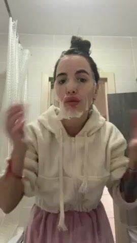 The video shows a woman in a bathroom applying a sheet mask to her face. She is wearing a white hoodie, a pink skirt, and her hair is in a bun. She has a tattoo on her left arm. She is smoothing the mask onto her face with her fingers. The bathroom has a shower curtain, a cabinet, and white tiles.