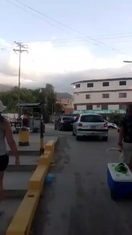 The video shows a street scene with several people and vehicles. In the foreground, a person wearing a white hat and a grey tank top walks away from the camera. To the right, a blue and white cooler on wheels is being pulled by an unseen person. Further down the street, a silver car is parked, and a black car is driving away. A multi-story building with the sign "HOTEL LUNA DEL MAR" is visible in the background. People are gathered around small stands or shops along the street. The environment appears to be a town or city with buildings and a street. The sky is overcast with some clouds.