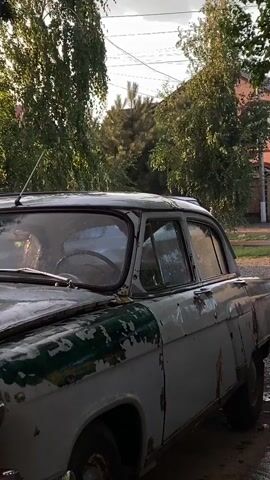 The video shows a close-up, side view of a white and green vintage car parked outdoors. The car appears weathered, with peeling paint and some rust visible. It is daytime, and the scene is framed by trees with green foliage. The sky is visible in the background, with some clouds. The camera is positioned at a low angle, focusing on the car's hood and side. The overall impression is one of an aged vehicle in a natural, somewhat overgrown setting.