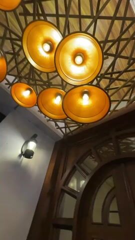 The video shows an ornate wooden door with glass panels. Above the door is a ceiling fixture with multiple round, orange-toned lights arranged in a pattern. The ceiling itself has a decorative wooden lattice design. To the left of the door is a gray wall with a small black light fixture. The camera angle is looking upwards towards the door and ceiling.  The door appears to be a grand entrance to a building or establishment, judging by its design. There are no people visible in the video.