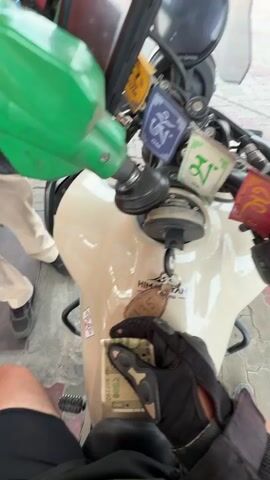 The video shows a person filling a motorcycle with fuel at a gas station in Taraori, India. The motorcycle is a Royal Enfield Himalayan, beige in color, and has prayer flags attached to the handlebars. The person is wearing a black riding glove and holding Indian Rupees. A gas station attendant in an orange shirt is operating the fuel pump. The gas pump displays "INDIANOIL" and has options for "DIESEL" and "PETROL". In the background, there are other vehicles, including an auto-rickshaw and a bus, as well as the gas station building and surrounding area. The weather appears to be overcast. The overall scene depicts a routine refueling stop during a motorcycle journey.