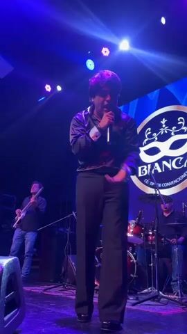 A male singer stands center stage, holding a microphone and singing. He wears a black collared shirt and black pants. To his left, a man plays a bass guitar. To his right, a drummer is seated behind a drum kit. A large circular logo featuring a mask and the word "BIANCA" is visible on a backdrop behind the performers. The stage is lit with blue and purple lights.