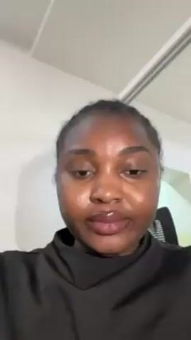 A medium shot shows a woman from the chest up, looking directly into the camera. She is in a room with a white ceiling. The woman has dark skin and her hair is braided back from her face. She is wearing a high-collared black shirt. The room appears to be an office, as an office chair is partially visible behind the woman. The lighting is bright and even.