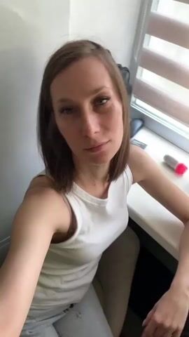 The video begins with a medium shot of a woman sitting at a desk in front of a window. She is looking directly at the camera with a neutral expression. Her hair is medium brown and shoulder-length. She is wearing a white tank top and light-colored pants. The background shows a window with blinds, a desk with a few objects on it, and a wall. The lighting is natural, coming from the window. In the next few frames, the woman brings her right hand up to her face and makes a peace sign with her fingers. She then lowers her hand and makes a gesture with her finger as if pointing. Finally, she holds the peace sign up again before the video ends. The overall impression is a casual, personal video, possibly a selfie or a short message.