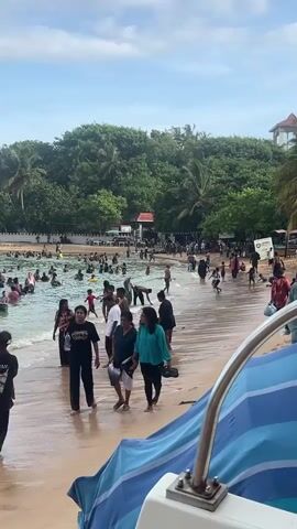 The scene is a crowded beach in what appears to be a tropical or subtropical location. Numerous people are in the water, some swimming and playing, while others are walking along the shoreline. The beach is sandy, and the water is relatively calm, with small waves lapping at the shore. There are trees and vegetation behind the beach, suggesting a natural environment. There are also boats visible near the shoreline. The sky is partly cloudy.