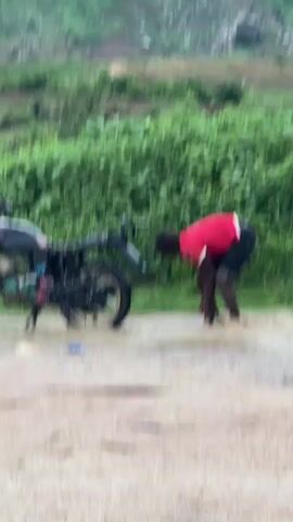 A person in a red shirt and dark shorts is washing the rear tire of a black motorcycle. The motorcycle is partially submerged in murky water on a dirt road. Lush green vegetation is visible behind the motorcycle, and a hilly, vegetated landscape is in the background. The water appears to be flowing across the road.