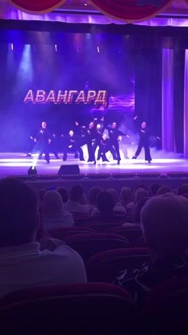 A video shows a group of performers on stage, illuminated by purple lights and stage fog, with a large screen displaying the word "АВАНГАРД". Several performers are on stage, wearing dark attire and moving rhythmically to music, some in a crouching position. The audience is visible in the foreground, seated in red chairs, observing the performance. The overall setting appears to be an auditorium or theater.