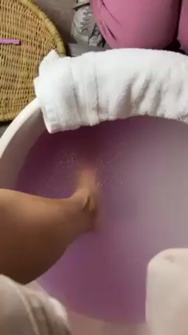 The video shows a person dipping their foot into a foot bath. The foot bath is a white plastic container filled with purple water that has a shimmery gold tint. A white towel is draped over the edge of the container. The person's leg is visible, and they are wearing a cream-colored outfit. In the background, there is a wicker basket, a purple nail file, a package of foot soak, and another person wearing pink pants. The floor is a light brown wood or wood-like surface. The camera is positioned directly above the foot bath, looking down.