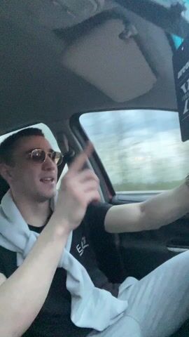 Two young men are seen inside a car, driving in what appears to be Seltsi, Bulgaria. The driver, wearing sunglasses, a black t-shirt, and a light grey sweatshirt tied around his shoulders, is singing along to music and smiling broadly. An "areon" car air freshener hangs from the rearview mirror. The camera, held by the passenger, captures a brief, blurry shot of the passenger themselves, wearing a pink and green shirt, before refocusing on the driver. Both individuals are laughing enthusiastically, conveying a joyful and energetic atmosphere. The scene unfolds during the daytime, with blurred scenery outside the car windows indicating motion.