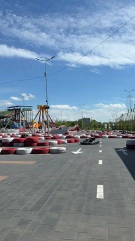 The video shows a go-kart track on a sunny day. There are go-karts on the track, and one is in motion. In the background, there are amusement park rides, including a Ferris wheel and a roller coaster. The sky is blue with some white clouds. The track is made of gray pavement and is lined with red and white tires. There are also some streetlights along the track.
