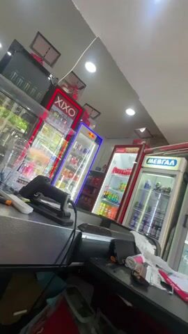 The video shows the interior of a store, likely a convenience store or mini-mart. The perspective is from behind a counter, looking towards several refrigerated display cases filled with beverages. A black barcode scanner is positioned on the counter. The display cases are brightly lit and stocked with various bottled and canned drinks, including brands like Red Bull. Some of the display cases have illuminated signage, such as "XIXO" and "MEBTAA". The overall environment appears to be a retail setting, with a focus on chilled beverages. The lighting suggests an indoor environment, possibly during the day or under artificial lights. There are no visible people in the frame.