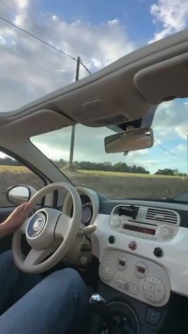 The video shows the interior of a white Fiat 500 car. The camera is positioned on the passenger side, looking towards the driver and the dashboard. The driver's hands are visible on the steering wheel, and their legs are visible in jeans. The dashboard features a cream-colored steering wheel with the Fiat logo in the center, a digital speedometer, a radio with buttons, and climate control dials. The car has a sunroof that is open, revealing a blue sky with white clouds. Outside the car, a rural landscape of fields and trees is visible under the sky. The car appears to be in motion, suggested by the slight blur of the exterior scenery and the position of the driver's hands on the wheel. A power pole is visible to the left of the car.