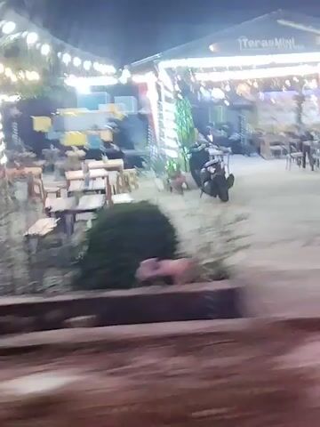 The video shows a nighttime scene with blurred motion. The first frame shows an outdoor seating area with tables and chairs. Lights are strung overhead, and a building with a stage is visible in the background. In subsequent frames, the view shifts to street-level, with cars, a scooter, and various buildings passing by. The overall impression is that of a moving vehicle capturing a quick glimpse of urban and possibly commercial areas at night. The motion blur makes it difficult to discern specific details, but the presence of outdoor seating and lighting suggests a lively atmosphere.