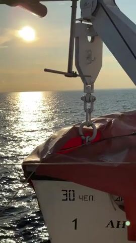 A close-up shot of a lifeboat is captured from the side, angled slightly upward. The lifeboat is predominantly white with an orange cover, and the text "30 ЧЕП." and the number "1" are visible on its hull. A davit, a piece of equipment used to launch lifeboats, is extended over the lifeboat, with a hook and chain mechanism holding it in place. The background features a calm sea bathed in the warm, golden light of the setting sun, which casts a shimmering reflection on the water's surface. The sky is partly cloudy, with the sun partially obscured by a cloud. The overall impression is one of tranquility and preparedness at sea during sunset.
