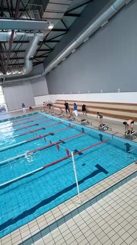 The video depicts an indoor swimming pool with several swimmers preparing for a race. The pool has multiple lanes divided by floating lane lines with red markers. Each lane has a starting block where swimmers are positioned, bending down and getting ready to dive in. The pool's tiles are a light blue color, and the water appears clean. Behind the starting blocks, a wall is marked with lane numbers from one to six. A person in white stands to the side, likely an official or coach. The environment is a well-lit, indoor facility with visible ventilation pipes and an industrial ceiling structure. The scene suggests a swimming competition or practice session.