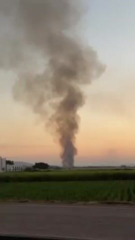 The video shows a thick column of gray smoke rising in the distance, against a backdrop of an orange and yellow sky, likely at sunset or sunrise. The smoke appears to be coming from beyond a green field and some trees. As the video continues, modern buildings are visible in front of the smoke plume. The camera is moving.