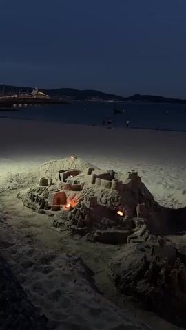 A sandcastle is illuminated by several small fires on a beach at night. The sandcastle is intricate, with turrets, archways, and walls. The fires are strategically placed within the structure, casting an orange glow and creating shadows. In the background, the dark ocean stretches out to the horizon, with a few distant lights from buildings or boats visible along the coastline. The sky is dark, indicating nighttime. A few people are faintly visible walking on the beach in the distance, silhouetted against the slightly lighter sky near the water. The overall scene is serene and somewhat magical, combining the art of sand sculpting with the drama of firelight against the night sky.