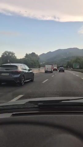 The video shows a first-person perspective from inside a vehicle traveling on a highway. The vehicle is in the right lane, with other cars and vans in front and to the left. The highway is bordered by a concrete barrier and trees. In the distance, mountains are visible under a cloudy sky. The lighting suggests it is either early morning or late afternoon. No individuals are clearly visible within the other vehicles. The focus remains on the road ahead and the surrounding environment.