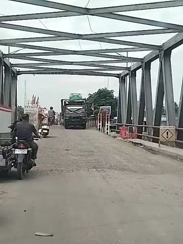 The video shows a bridge with a metal framework overhead. The bridge is made of cement. A black truck is driving towards the camera. A motorcycle with a sidecar is on the left of the frame, and another motorcycle is farther ahead. There is a warning sign on the right side of the bridge. There are buildings and trees visible in the background. The sky is overcast. The road surface appears uneven and slightly damaged.