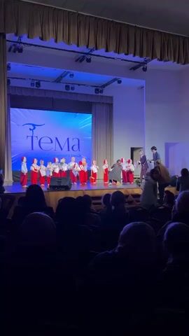 A stage performance is taking place indoors. A group of approximately 15 children dressed in red and white folk costumes are lined up on a stage. Some hold bouquets. The stage has a large screen displaying a logo and text in Russian. A man and woman in casual clothes are walking on the stage, possibly distributing flowers or congratulating the children. An audience sits in the dark, observing the performance. The lighting on stage is predominantly blue. The stage is decorated with beige curtains.